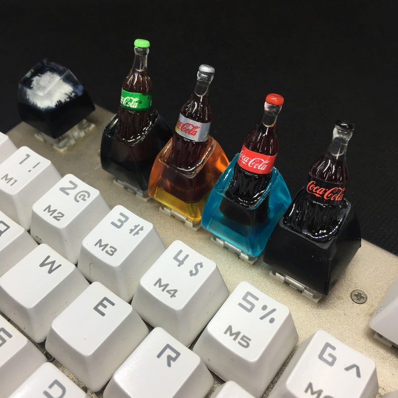 May include: Four custom mechanical keyboard keycaps featuring miniature Coca-Cola bottles embedded in translucent resin bases of black, amber, cyan, and black. The bottles have distinct labels and caps, positioned above white keys with black alphanumeric characters.