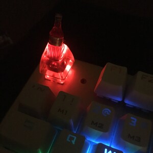 May include: A translucent, red-lit custom keycap shaped like a small glass bottle, installed on a white mechanical keyboard. The keycap glows with a bright red light, contrasting with the white keys labeled with numbers and M-series markings.