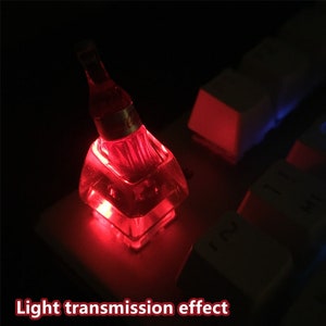 May include: A translucent red mechanical keyboard keycap shaped like a miniature glass bottle with the Coca-Cola logo. The keycap is illuminated from within, casting a bright red glow across the surrounding white keyboard keys in a dark setting.