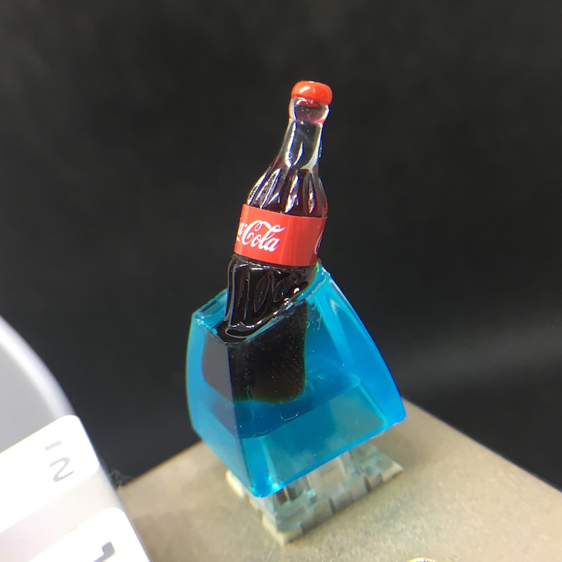 May include: A custom mechanical keyboard keycap featuring a miniature dark brown glass bottle with a red label that reads Coca-Cola, set inside a translucent bright blue resin block. The bottle has a red cap and a contoured shape.