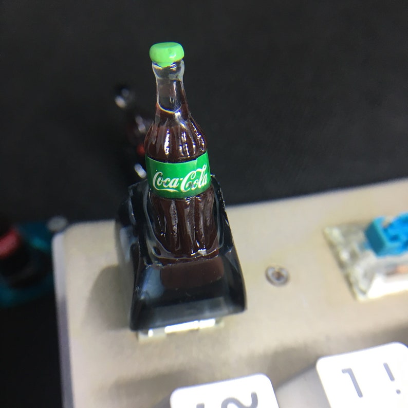 May include: A miniature Coca-Cola bottle keycap featuring a dark brown liquid, a green label with white Coca-Cola text, and a light green cap. The bottle is mounted on a black base, designed to fit onto a mechanical keyboard switch.