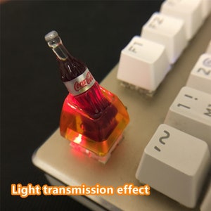 May include: A custom mechanical keyboard keycap featuring a miniature Coca-Cola bottle design. The keycap is made of translucent orange resin, allowing internal red light to shine through. The text Light transmission effect is visible.