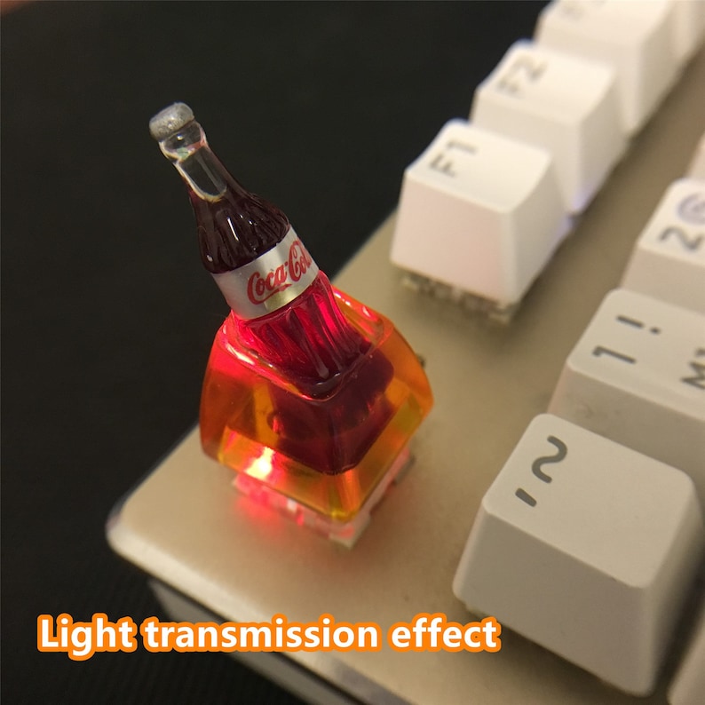 May include: A custom mechanical keyboard keycap featuring a miniature Coca-Cola bottle design. The keycap is made of translucent orange resin, allowing internal red light to shine through. The text Light transmission effect is visible.