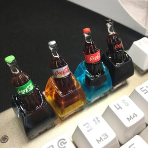 May include: Four custom mechanical keyboard keycaps featuring miniature Coca-Cola bottles embedded in translucent resin bases of black, orange, blue, and black. Each bottle has a distinct colored cap and the Coca-Cola logo printed on the label.