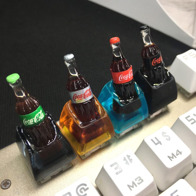 May include: Four custom mechanical keyboard keycaps featuring miniature Coca-Cola bottles embedded in translucent resin bases of black, orange, blue, and black. Each bottle has a distinct colored cap and the Coca-Cola logo printed on the label.