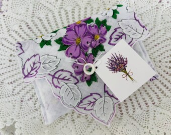 May include: White handkerchief with purple floral and leaf design. The handkerchief has a scalloped edge and a small tag with a lavender bouquet illustration. A white button is attached to the handkerchief.