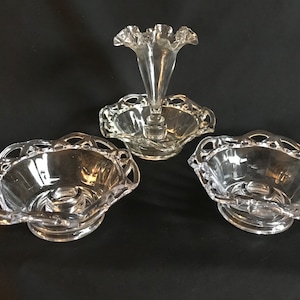 May include: A clear glass three-piece centerpiece with a central vase and two bowls. The vase has a scalloped rim and the bowls have a scalloped edge.