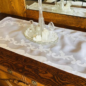 May include: A white, rectangular table runner with an embroidered floral design. The runner is placed on a dark wooden surface, with a glass vanity set and mirror in the background. The vanity set includes perfume bottles and a decorative tray.