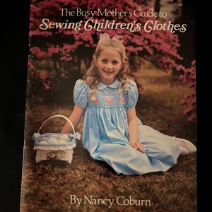 May include: A book cover titled "The Busy Mother's Guide to Sewing Children's Clothes" featuring a young girl in a blue dress. The girl is smiling and holding a basket with eggs. The author's name, Nancy Coburn, is at the bottom.