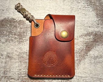 May include: A brown leather card holder with a brass snap closure, a metal eyelet, and a paracord lanyard featuring a brass bead. The front is embossed with a circular mountain logo and the text Night Shift Leather, featuring visible stitching.