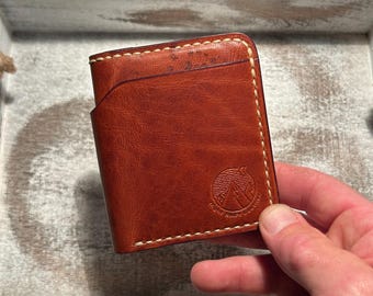 May include: A brown leather bifold wallet featuring a curved card slot opening and light-coloured contrast stitching along the edges. The front surface displays a circular embossed logo depicting a mountain range under a crescent moon and stars.