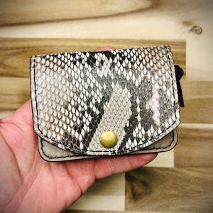 May include: A small, brown and white snakeskin wallet with a gold button closure. The wallet is held in a hand.