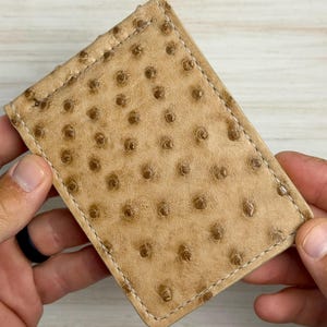 May include: A rectangular tan wallet featuring a distinct textured pattern of small, raised circular bumps across the surface. The wallet is constructed with visible white stitching along the edges, creating a clean, structured border around the perimeter.