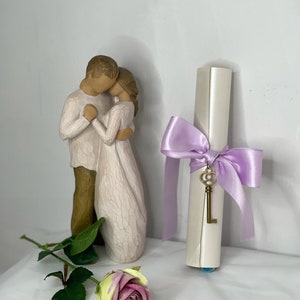 May include: A cream-coloured scroll tied with a lavender ribbon and a gold key charm. A figurine of a couple embracing and a pink and yellow rose are also present. The items are arranged on a white surface.