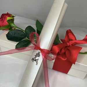 May include: A white scroll tied with a red ribbon and a silver reindeer charm. The scroll is sitting on top of a white box with a red satin ribbon.