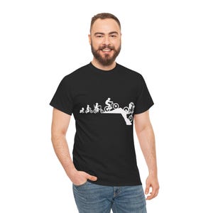 May include: Black t-shirt featuring a white graphic depicting the evolution of a cyclist, from a toddler on a balance bike to a person performing a stunt. The design shows a progression of bicycle types and rider skills.