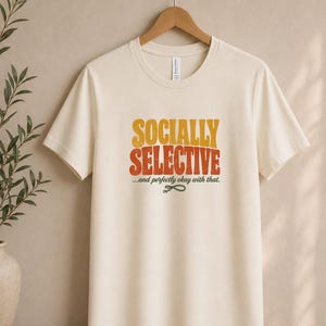 May include: A cream-colored short-sleeve t-shirt featuring the text "SOCIALLY SELECTIVE" in large, distressed yellow and orange letters, with the smaller text "...and perfectly okay with that." and a small dark green graphic icon below it.