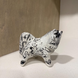 May include: A ceramic figurine of a dog, painted white with black decorative patterns. The dog is depicted in a seated position with its head up. The figurine has three legs and is set against a neutral background.