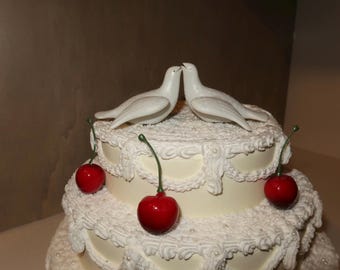 May include: A tiered wedding cake with white frosting and decorative icing. Two white dove figurines are perched on top, with three red cherries as accents. The cake has a classic, elegant design.