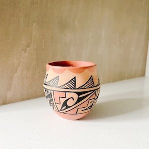 May include: A small, hand-painted ceramic pot with a geometric pattern in black and pink. The pot is made in the Southwestern style.