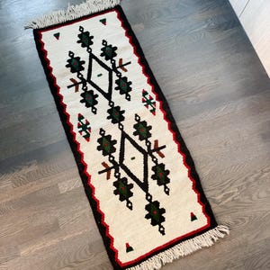 May include: A rectangular, woven rug with a cream-colored background and a central geometric design in black, green, and brown. The rug has a black border with a red trim and white fringe on the short ends. The rug measures approximately 36 inches long.