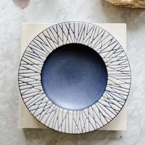 May include: A round ceramic plate with a navy blue center and a cream-colored, textured rim. The rim features a geometric pattern of intersecting lines. The plate is set on a light-colored surface, suggesting a decorative or serving piece.