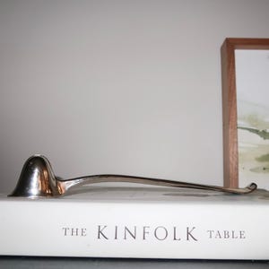 May include: A silver-colored ladle with a rounded bowl and a long, curved handle. The ladle rests on a white book titled "The Kinfolk Table". The background includes a framed picture and a neutral-colored wall.