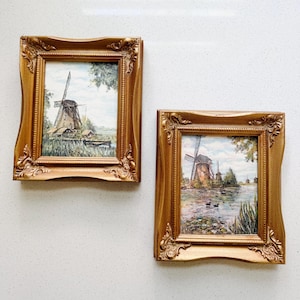 May include: Two rectangular wall art pieces featuring paintings of windmills in ornate gold frames. The left painting depicts a windmill in a field, while the right painting shows a windmill near a body of water with two ducks swimming in the foreground.