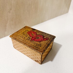 May include: A rectangular wooden jewelry box with a floral design and the word "Jewels" on the lid. The box has a woven pattern on the sides and a red and pink floral design on the top.