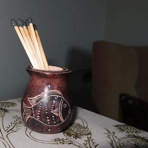 May include: A small, brown ceramic vase with a fish design etched on the side. The vase is filled with several wooden matches with black tips. The vase sits on a patterned tablecloth.