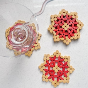 May include: Three decorative coasters with a starburst design. The coasters are made of natural-colored wood and red wooden accents. A glass is being filled with a clear liquid, resting on one of the coasters.