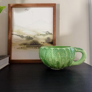 May include: A light green ceramic mug featuring a textured, embossed pattern resembling overlapping leaves or scales on its exterior surface. The mug has a rounded bowl shape and a curved handle extending from the right side of the vessel.