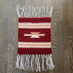 May include: A rectangular, handwoven rug with a deep red background. The rug features a central geometric design in cream and tan, with horizontal stripes and white fringe on the top and bottom. The rug is approximately 12 inches wide and 18 inches long.