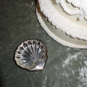 May include: A small, silver-colored seashell-shaped dish with a detailed, ridged design. The dish is set on a green surface, with a portion of a white, ornate, decorative object in the background.