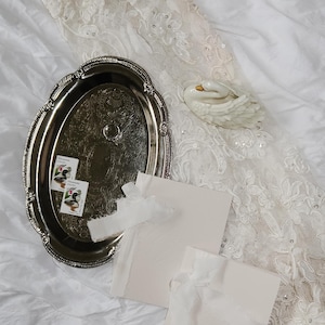 May include: A silver oval tray with ornate edges holds two vintage stamps, a ring, and two cream-colored cards tied with white ribbon. A white ceramic swan figurine rests on a white lace fabric.