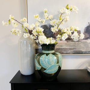 May include: A green ceramic vase with a leaf design and white flowers in it. The vase is sitting on a black table.