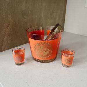 May include: A set of three orange glass drinkware with gold sun designs. The set includes a large ice bucket and two shot glasses.