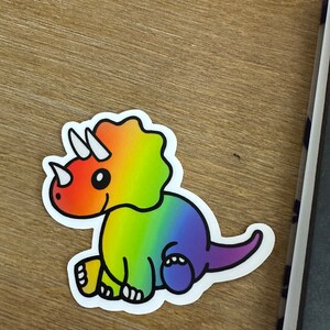 May include: A rainbow-colored cartoon triceratops sticker. The dinosaur is sitting down with its tail curled behind it.