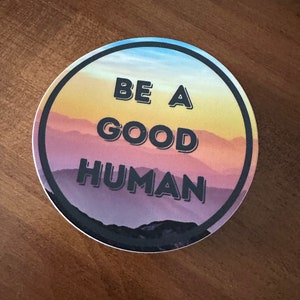 May include: A round sticker with a sunset mountain scene and the text "Be a good human" in black.