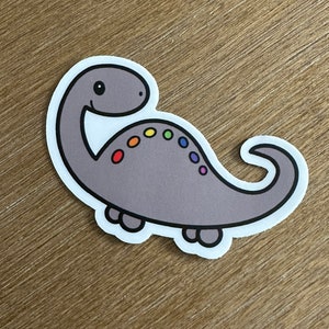 May include: A gray cartoon dinosaur sticker with a rainbow pattern on its back. The dinosaur has a smiling face and is standing on four legs.