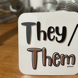 May include: A square white sticker with the words "They/ Them" in black and brown bubble letters. The sticker has a rounded edge and the text is in a playful font. The sticker is on a wooden surface.