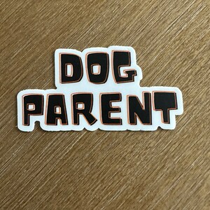 May include: A white sticker with the words "DOG PARENT" in black block letters with an orange outline. The sticker is on a light brown wooden surface.