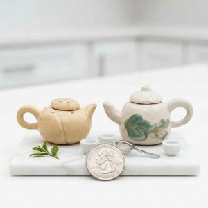 May include: Two miniature ceramic teapots, one beige with small dots on the lid and one off-white with a green leaf design, accompanied by three tiny white cups and a small metal strainer, with a US quarter coin for scale on a marble slab.