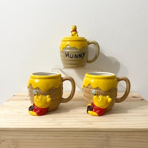 May include: Three ceramic mugs shaped like honey pots with yellow dripping glaze. Two mugs feature a sculpted yellow bear in a red shirt with a blue bee. The top mug has a lid with a small bear figurine and the word HUNNY printed in black.