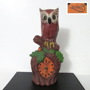 May include: A vintage owl-shaped clock, primarily in shades of red and brown, perched on a tree trunk base. The clock face is orange with black markings and a key. The name "DELORES" is visible on a small wooden plaque.