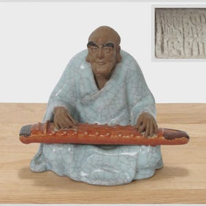 May include: A ceramic figurine of a seated person with brown skin and dark eyebrows, wearing a light blue robe with a crackle glaze finish. The figure holds a long, reddish-brown musical instrument across the lap. A square seal mark is visible.