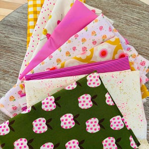 Puede incluir: Una colección de muestras de tela coloridas, con varios patrones. Los diseños incluyen flores rosas, cuadros amarillos, rayas rosas y tela blanca con motas rosas y doradas. Las telas están dispuestas en forma de abanico, mostrando la variedad de patrones y colores.