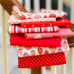 May include: A stack of various red and pink fabric pieces with strawberry patterns, polka dots, and stripes. The fabrics are folded and stacked, showcasing a variety of designs and textures. Ideal for crafting and sewing projects.