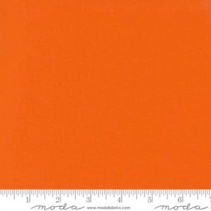 Può includere: Un campione di tessuto arancione brillante e solido con una trama tessuta. Nella parte inferiore è visibile un righello bianco che mostra le misure in centimetri da 1 a 18, con il testo "moda" e "www.modafabrics.com" stampato sul bordo.