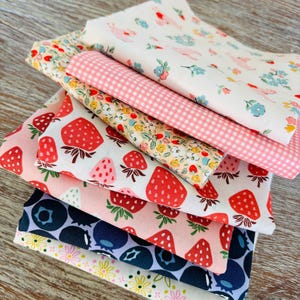 Peut inclure: Une pile de tissus imprimés avec des motifs variés : fraises, myrtilles, fleurs et carreaux vichy. Les tissus présentent un mélange de couleurs rose, rouge, bleu et jaune, parfaits pour le patchwork ou les travaux manuels.